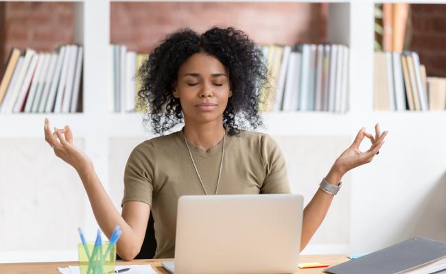 An African-American woman is meditating while sitting at work desk.