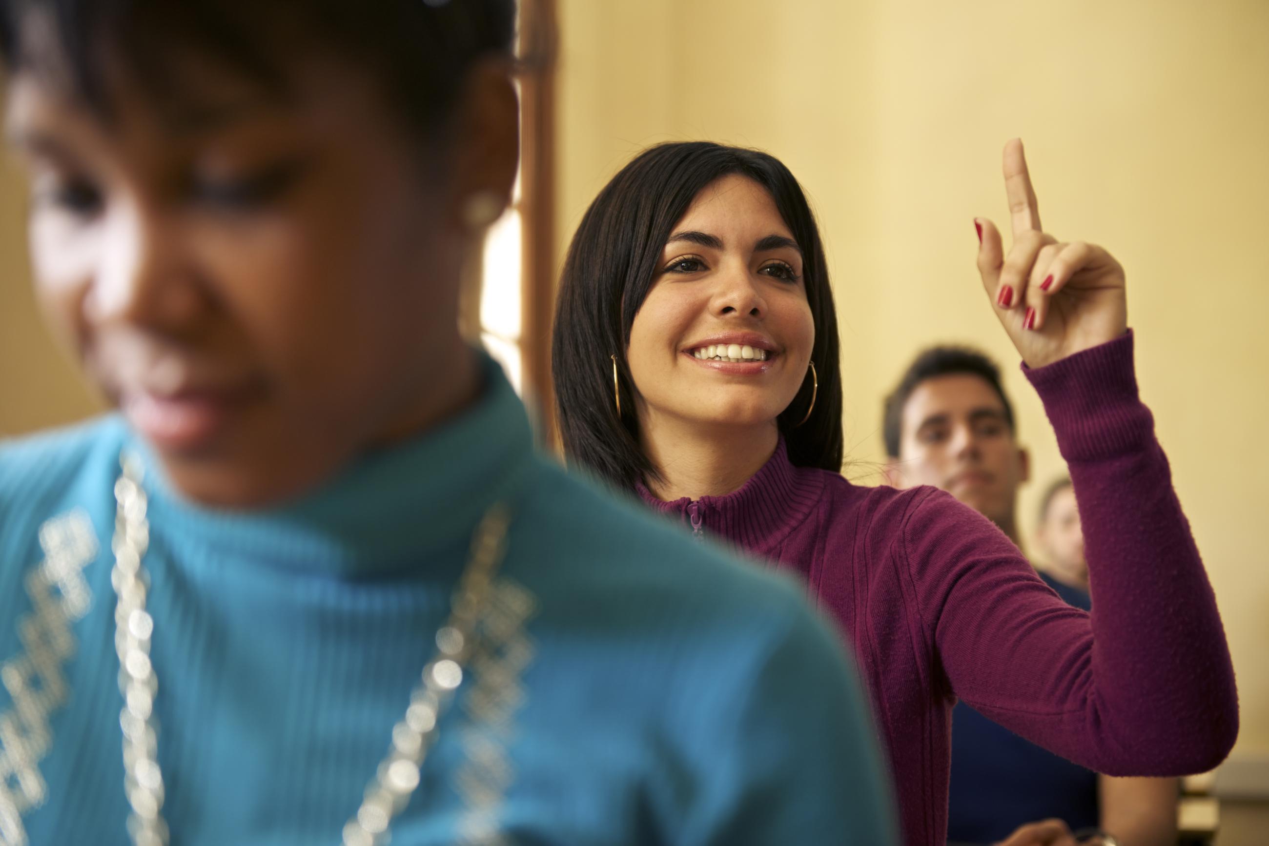 Students sitting in a classroom. One female student is raising her hand.